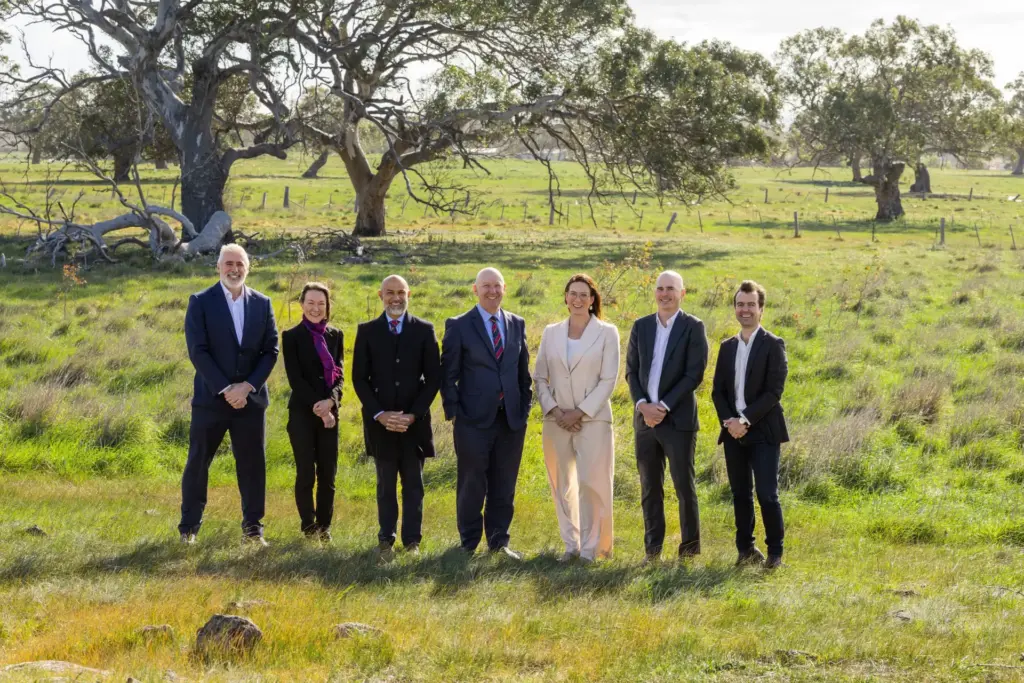 Seven people in business attire stand in a grassy field with scattered trees and a fence in the background, posing together and smiling on a sunny day.
