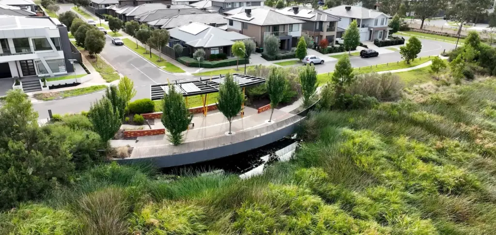Aerial view of a modern suburban neighborhood with houses, a small curved bridge over water, a shaded seating area with benches, and lush green vegetation surrounding the area.