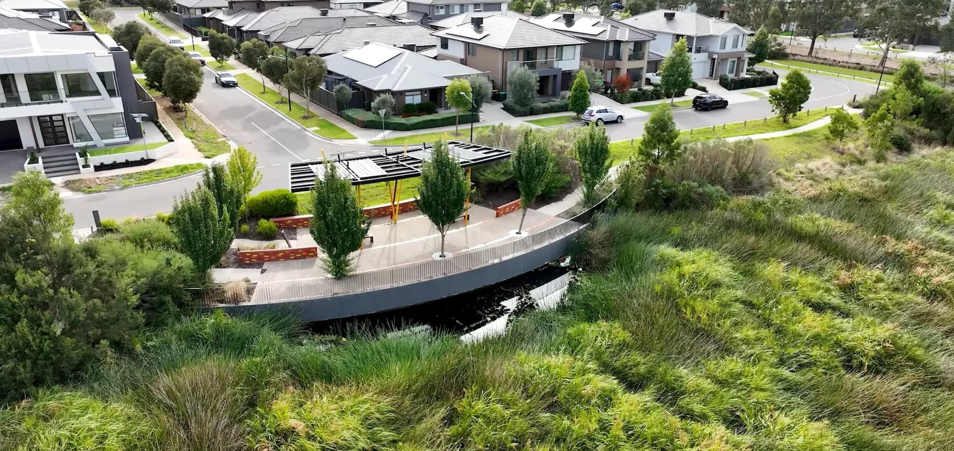 Aerial view of a modern suburban neighborhood with houses, a small curved bridge over water, a shaded seating area with benches, and lush green vegetation surrounding the area.