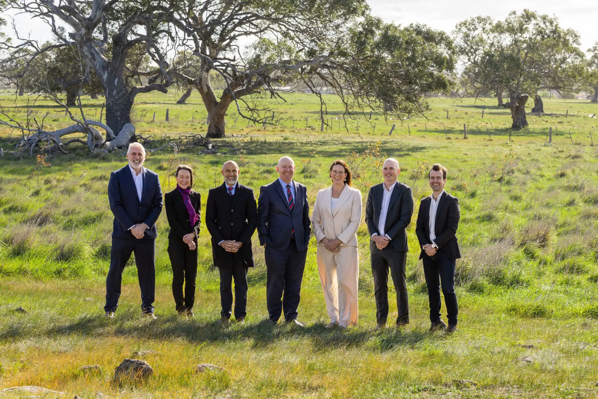 Seven people in business attire stand in a grassy field with scattered trees and a fence in the background, posing together and smiling on a sunny day.