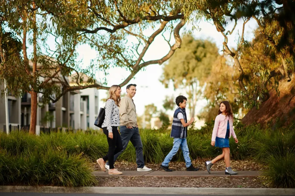 A family of four, two adults and two children, walk together outdoors on a pathway surrounded by greenery and trees, with modern buildings visible in the background.