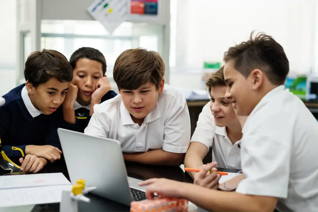 Five boys in school uniforms sit around a laptop, engaged and smiling, in a classroom setting. Notebooks and stationery are on the desk, and they appear to be collaborating on a project.