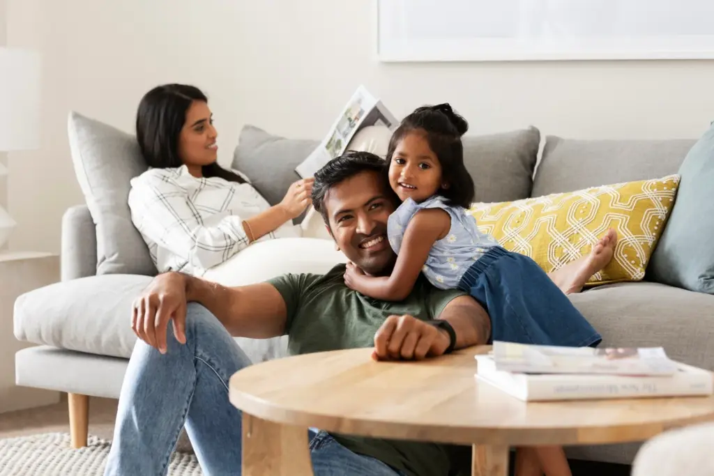 A man sits on the floor smiling as a young girl hugs him. Behind them, a woman relaxes on the sofa reading a magazine. The scene is cozy, with neutral-colored furniture and bright cushions.
