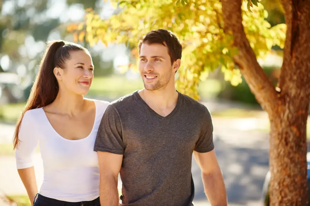 A smiling couple walks outdoors on a sunny day. The woman, with long brown hair in a ponytail and wearing a white top, looks at the man, who has short brown hair and wears a gray T-shirt. Trees with yellow-green leaves are nearby.